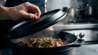 Chef lifting lid from cast iron skillet revealing steaming cooked meat with herbs in professional kitchen. Culinary cooking process with steam and seasoned dish.