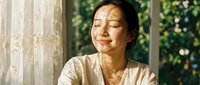 Peaceful Asian woman with closed eyes enjoying warm sunlight streaming through window with lace curtains, expressing serenity and mindfulness in cozy home interior.