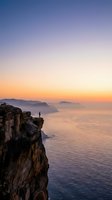 Solitary figure standing on rocky cliff edge at sunrise overlooking misty ocean waters and distant islands, conveying contemplation and adventure for travel and lifestyle editorial use.
