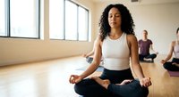 Young African American woman practicing meditation in lotus pose during yoga class in bright studio with natural light and wooden floors.