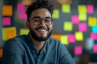Happy young mixed-race businessman with glasses and beard smiling at camera in modern office with colorful sticky notes background for creative workspace.