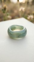 Polished jade ring with soft green and white marbling, displayed on white surface with blurred floral background. Suitable for jewelry, luxury goods, and lifestyle marketing.