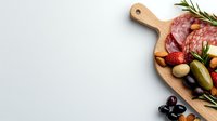 Wooden cutting board with assorted antipasto ingredients including salami, olives, pickles, and herbs on light background with copy space.