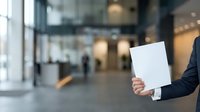 Businessman in dark suit holding blank white paper sheet in modern office lobby with blurred background, corporate communication and business messaging concept.