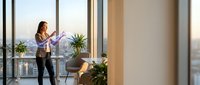 Young Asian businesswoman using smartphone in modern office with floor-to-ceiling windows overlooking city skyline during golden hour for professional communication.