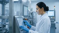 Female scientist in white lab coat operating industrial equipment control panel in modern pharmaceutical manufacturing facility.
