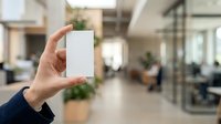 Person holding blank white business card in modern office environment with blurred colleagues working in background, suitable for corporate branding and marketing materials.