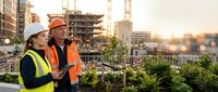 Two male construction workers in safety gear inspect rooftop garden with city skyline and cranes at sunset, discussing sustainable urban development and green building practices.