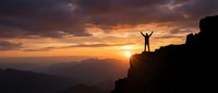 Silhouette of victorious hiker with raised arms standing on mountain peak at golden sunset with dramatic sky and layered mountain ranges in background.