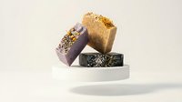Artisan handmade soap bars with natural ingredients and herbs stacked on white platform against neutral background for spa and wellness products.