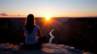 Woman meditating at sunrise on canyon edge with dramatic sunburst and valley views. Peaceful morning yoga practice in nature for wellness and mindfulness.