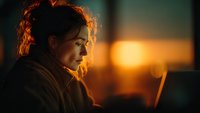 Young woman working on laptop at sunset with warm golden light creating atmospheric silhouette for remote work and digital lifestyle concepts.