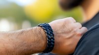 Close up view of male wrist wearing black braided leather bracelet against blurred natural background, showing modern casual fashion accessory and jewelry styling.