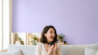 Young woman with long dark wavy hair and olive complexion, wearing casual beige shirt with black choker, sitting on cream sofa against purple wall, smiling joyfully while looking to side.