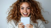 Young woman with curly hair and bright smile holding smartphone, wearing white knit sweater against light background. Joyful expression and natural look.