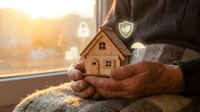 Hands holding wooden house model with security icons floating above, representing home protection, insurance, and real estate investment concepts.