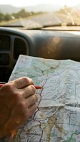 Driver holding map with pencil while planning route during road trip, vehicle dashboard and mountain landscape visible through windshield in background.