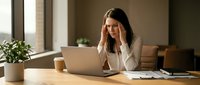 Stressed young businesswoman with headache working on laptop at office desk, feeling overwhelmed by workload and deadlines in modern workplace.