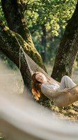 Woman with long reddish-brown hair relaxing in hammock strung between moss-covered trees in forest setting, wearing casual light clothing, peaceful outdoor lifestyle scene.