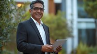 Smiling mature South Asian businessman in formal suit using digital tablet outdoors near modern office building with greenery.
