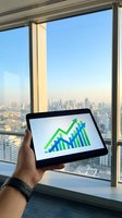 Businessman holding tablet displaying green and blue growth chart with cityscape skyline visible through office window behind.