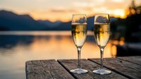 Two champagne flutes filled with sparkling wine on wooden dock at sunset over mountain lake creating romantic celebration atmosphere.