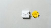 Small white gift box with silver ribbon bow next to bright yellow daisy flower on neutral gray background for greeting cards and celebrations.