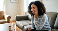 Young African American woman experiencing toothache pain sitting on couch at home holding jaw with hand showing dental discomfort and oral health issues.