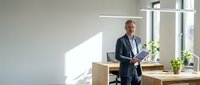 Confident mature businessman in suit standing in modern office workspace with natural lighting and wooden furniture for corporate presentations.