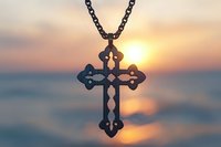 Ornate metal cross pendant on chain silhouetted against golden sunset over water, symbolizing faith, spirituality, and peaceful contemplation.