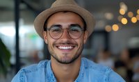 Young Hispanic man wearing fedora hat and glasses smiling confidently in modern office setting with bokeh lights background. Casual professional portrait.