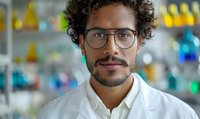 Mixed race male scientist with curly hair and glasses wearing white lab coat smiling confidently in research laboratory with colorful glass containers in background.