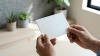 Hands holding blank white card with potted plants and window in background, suitable for business branding, greeting card design, and marketing mockups.