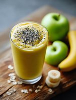 Fresh fruit smoothie with chia seeds in glass, surrounded by green apples, bananas and oats on wooden board, perfect for healthy breakfast.