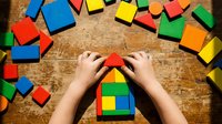 Child hands building colorful wooden block house on rustic table surrounded by scattered geometric toy blocks for creative play and learning development.