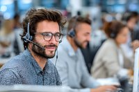 Smiling young male customer service representative with headset and glasses in busy call center environment.