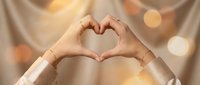 Two hands forming a heart shape gesture against a warm golden bokeh background, expressing love, care, and positive emotions for Valentine's Day or wellness concepts.