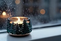 Glowing candle holder with decorative winter pine trees design on windowsill against snowy background with bokeh lights and raindrops on window glass.