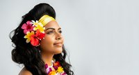Young woman with dark hair and South Asian ethnicity wearing vibrant multicolored flower headpiece and matching floral accessories, looking upward against white background for beauty and wellness campaigns.