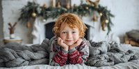 Cheerful child with curly hair wearing festive mittens, resting on cozy knitted blanket against holiday decorated fireplace with garland and lights.