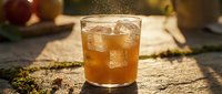 Refreshing iced apple cider cocktail in a glass on a rustic stone surface, golden sunlight, sparkling droplets, autumn mood.