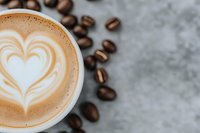 Creamy latte with heart-shaped foam art surrounded by scattered roasted coffee beans on gray concrete surface, close-up view with soft focus background.