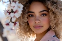 Young mixed race woman with natural curly blonde hair and glowing skin poses near blooming cherry blossoms in soft spring light, expressing serenity and natural beauty.