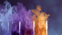 Colorful smoke rising from laboratory glass beakers with blue, purple, and orange chemical reactions creating dramatic atmospheric effect against dark background.