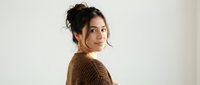 Young brunette woman in brown knit sweater looking over shoulder with confident expression against neutral background for lifestyle photography.