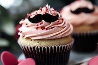 Pink frosted chocolate cupcake decorated with black mustache topper and red sprinkles on dark background, selective focus on creamy swirled buttercream.
