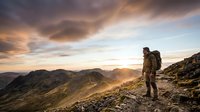 Male hiker with backpack standing on mountain ridge at sunset, overlooking vast valley landscape with golden light and dramatic clouds.