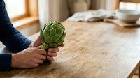 Fresh artichoke held in hands on wooden kitchen counter, ready for cooking and meal preparation.