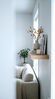 Minimalist living room corner with floating wooden shelf, dried pampas grass in ceramic vase, books, candle, beige sofa, knit throw blanket and potted plant.