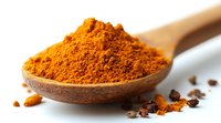 Ground turmeric powder in wooden spoon with scattered spice particles on white surface, bright orange aromatic seasoning for cooking and wellness uses.